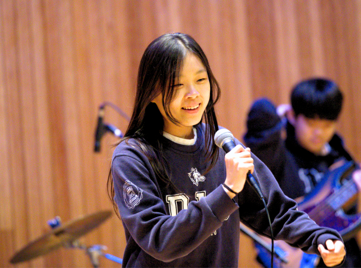 Stella Lee in seventh grade, the lead vocalist of her band, extends an inviting hand as if to urge the audience to join in along the fun. The secondary student body claps and whistles in response. 