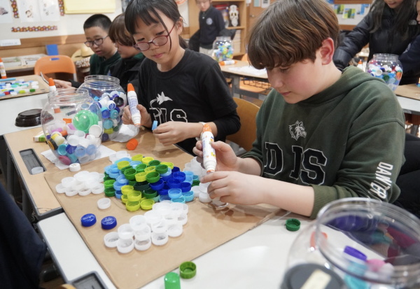 Fifth graders Aiden Hwang and Akari Kojima design their very own turtle. Hwang carefully glues the bottle caps onto the cardboard, adding final touches. Kojima lends him a hand. 