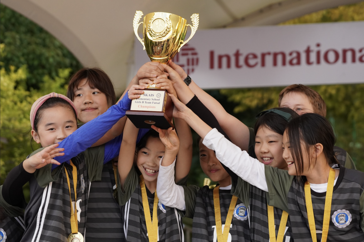 The Girls B team holds up their trophy proudly, medals around their necks. They giggle as they celebrate their well-deserved success. 