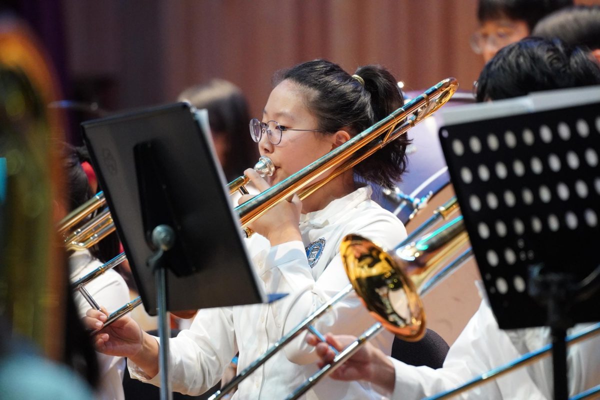 Eighth-grader Elba Jekal plays the trombone with focus and precision during an ensemble performance, showcasing dedication and skill. The backdrop of sheet music and brass instruments highlights the collaborative spirit of the orchestra. 