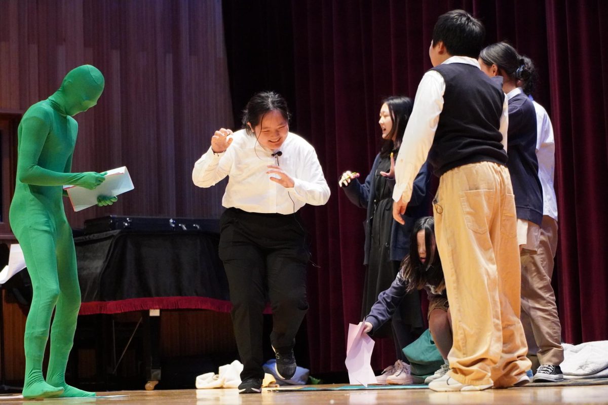 Eighth grader Katniss Kim, portraying Trunchbull, dashes off the stage in a dramatic escape after a surprise encounter with the mysterious green screen figure. The comical moment captivates the audience, leaving them in fits of laughter.