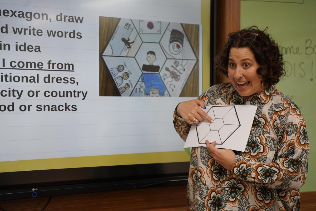 Mrs. Nunes’ eyes light up as she points at the empty hexagon template. Her curly hair bounces with her animated gesture, reflecting her excitement. 