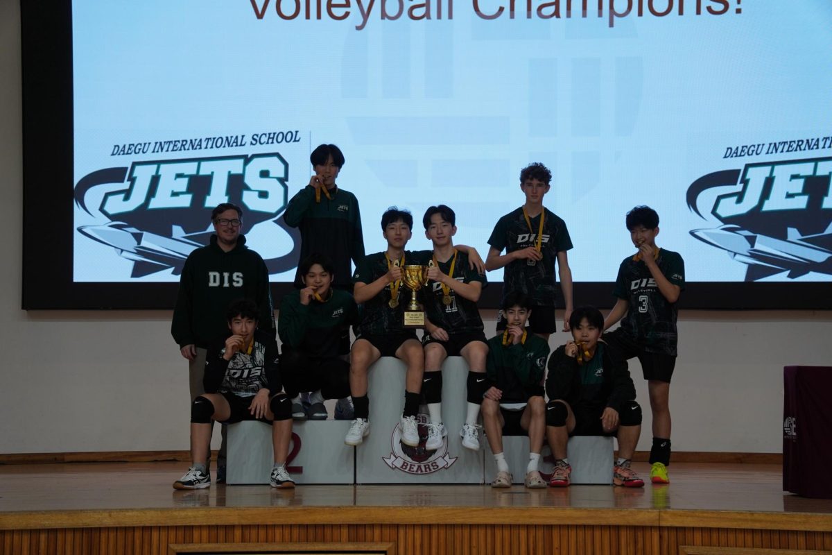 The varsity boys celebrate their four-peat trophy on the podium. Senior captains Brandon Hyun (11) and Chris Ryu (7) stand in the middle holding the trophy. To celebrate their success to the fullest, they bite their gold medals.