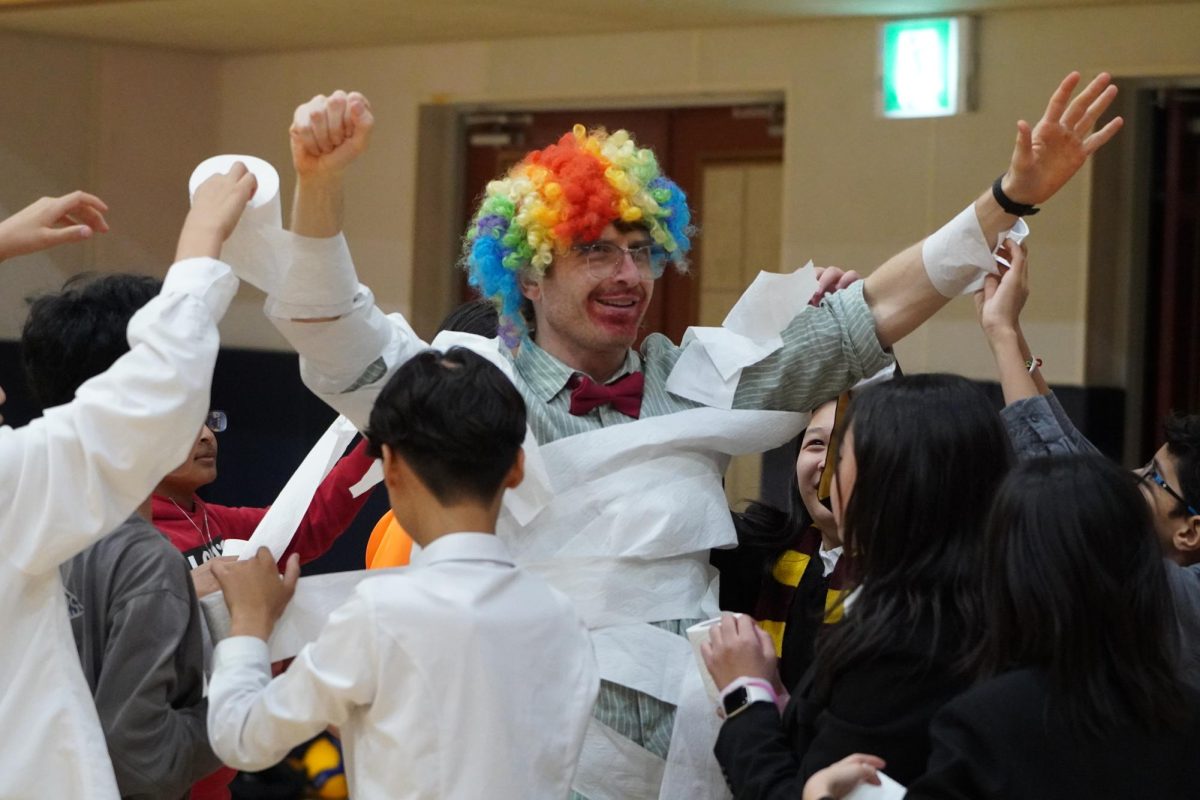 Swarmed by middle schoolers, Mr. Lipsky throws his hands up to halt the chaos for the mummy wrapping booth. As students cloak him in layers of toilet paper, even more rush in to join the frenzy.