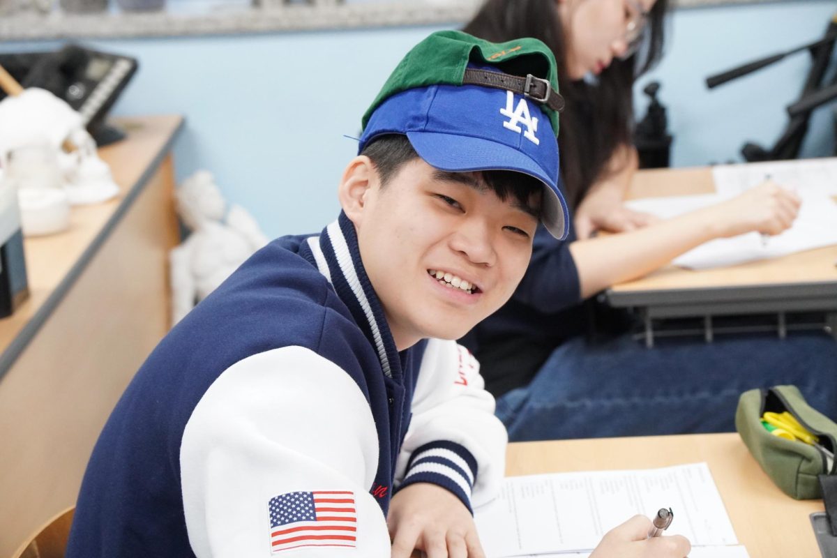 Sophomore Luke Cho doubles up on school spirit with blue and green caps. His goofiness brings out a chuckle from his classmates as they chug through their worksheets.