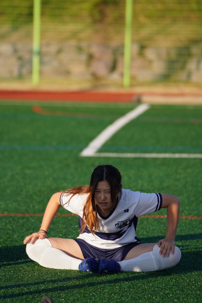 Left back Esther Kang (53) stretches on the grass during warm-ups, preparing for the match ahead. Focused and alert, she readies herself to stop any attack from breaking through the back line. 