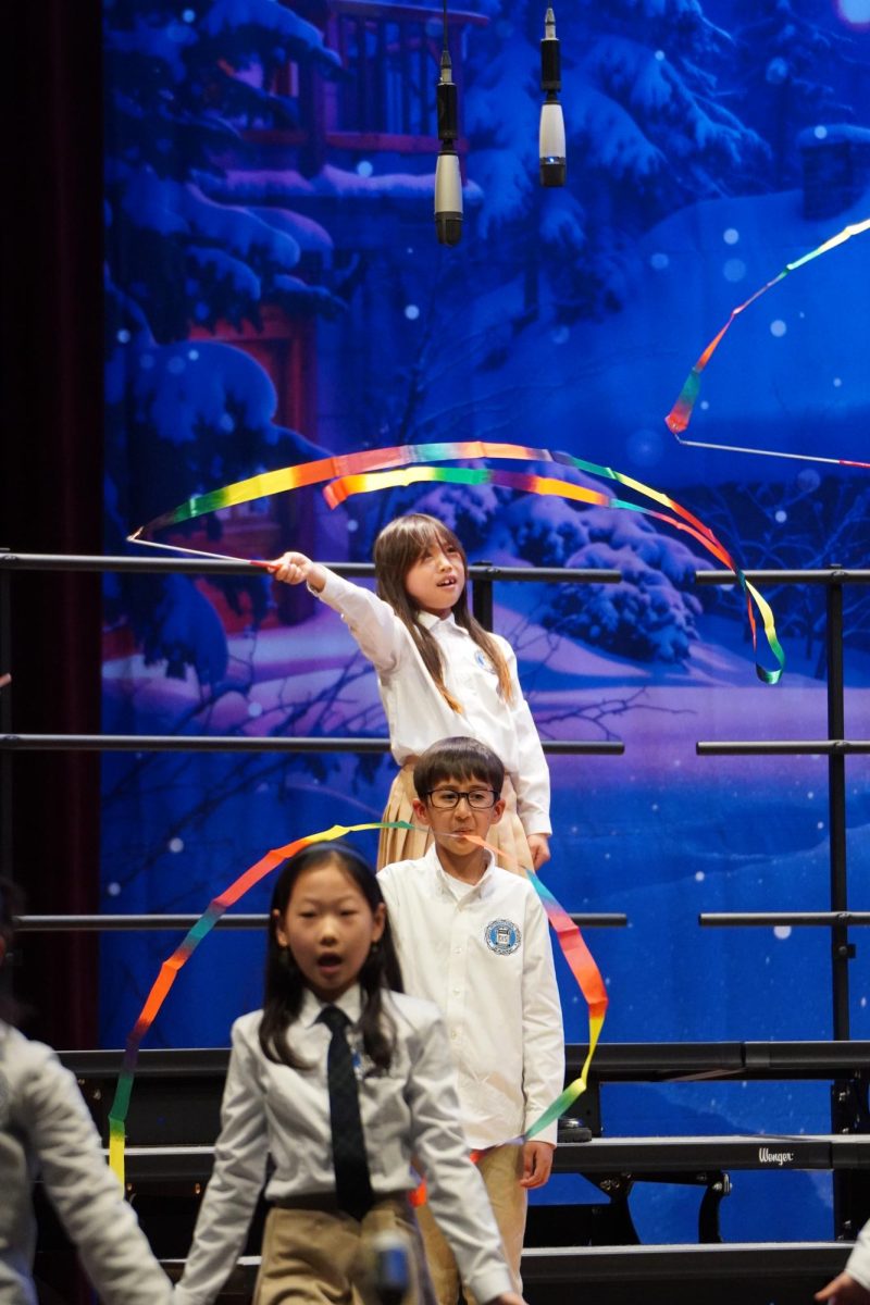 Selena Ahn in third grade waves the rainbow-colored ribbon sticks in wide, colorful motions. The vivid background and warm voices of students add a vibrant touch to the night.