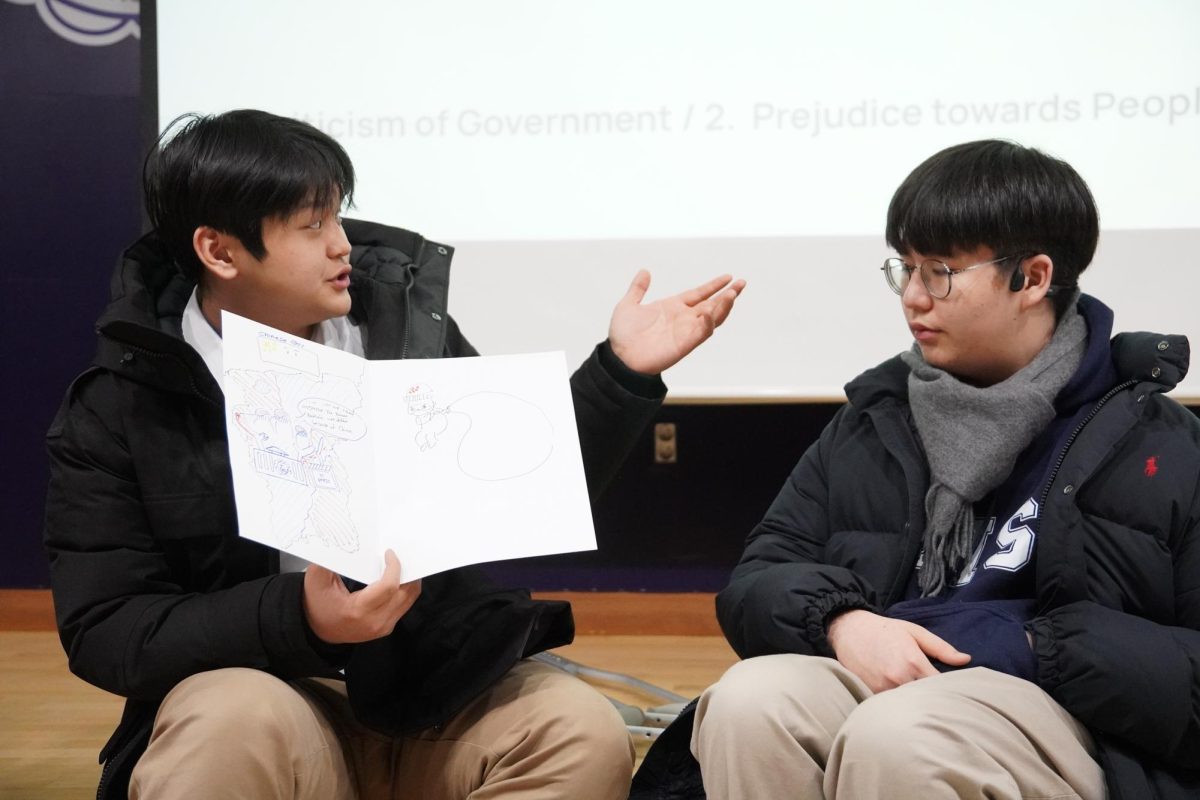 Junior Justin Son gestures toward a hand-drawn poster as he explains how political narratives shape public bias. Seated beside him, Jake Park listens closely, preparing to share his own response.