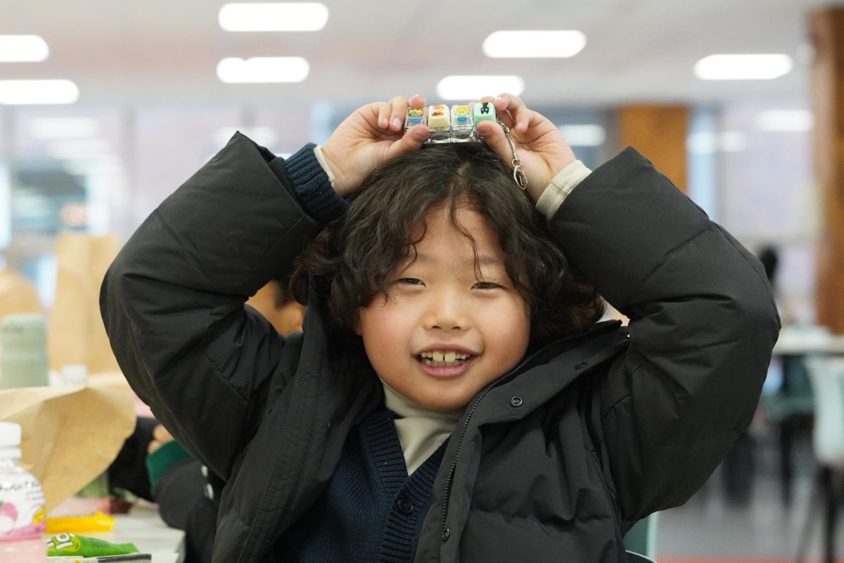 First grader Julian Jung proudly presents his craft during a creativity session in the cafeteria. The figures feature playful tiles and expressive, whimsical faces. 