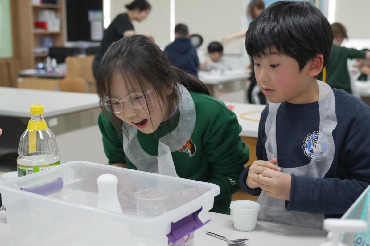 Third graders Yitong Li and Takaharu Watanabe lean over the table and watch their volcanoes erupt after they pour white vinegar. Yitong watches with her mouth open while Takaharu studies the reaction with his hands together. 