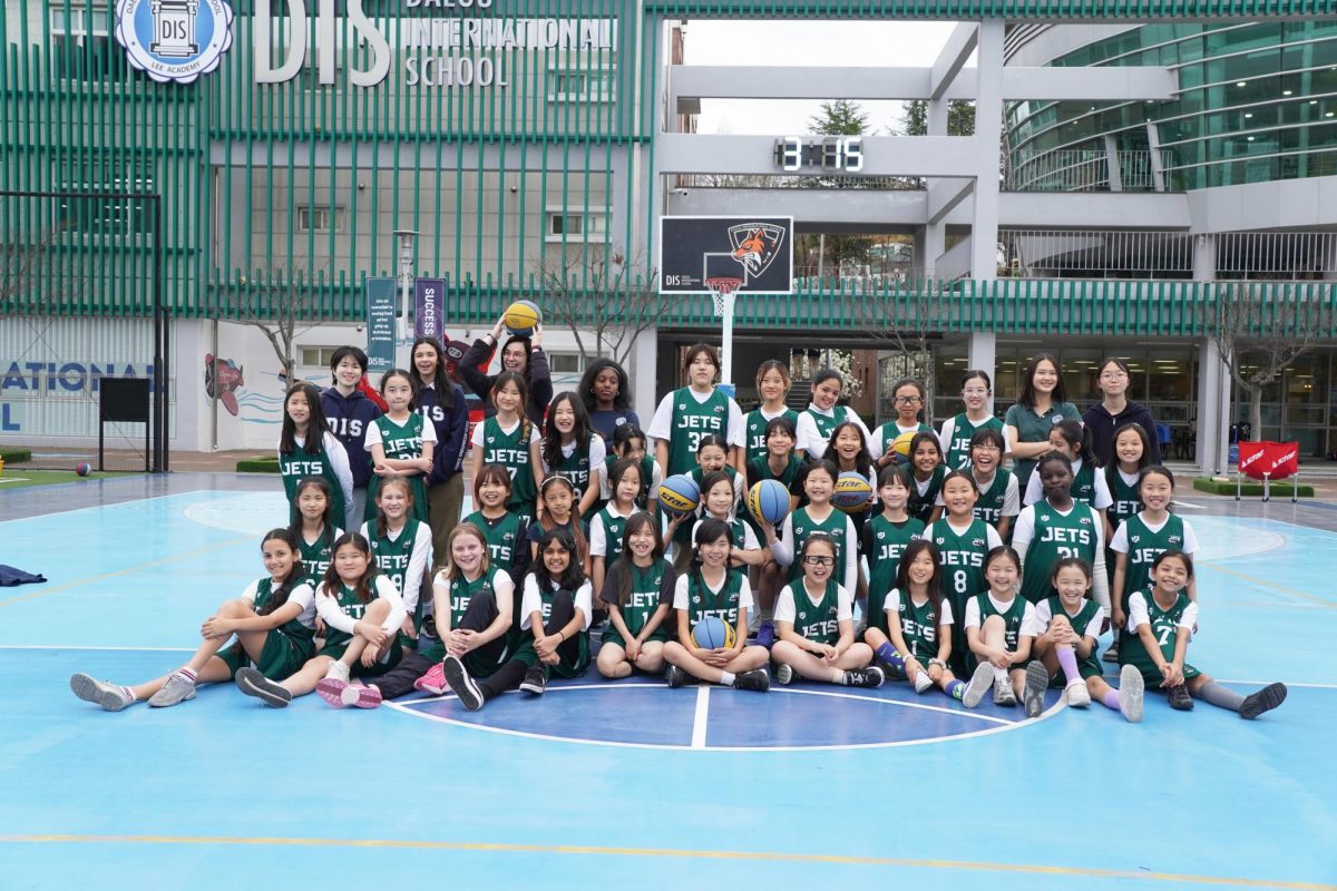 The elementary girls basketball team clutch their basketballs as they line up across the court to celebrate their successful season. They kneel down and raise the balls overhead as they represent DIS with confidence and energy heading into the season. 