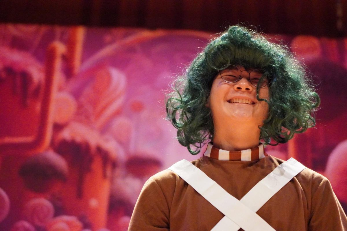 An Oompa-Loompa (Daniel Beck) runs on stage as Willy Wonka (Katniss Kim) barks out orders to clean up the mess the five Golden Ticket winners made. “Oompa loompa doompety doo!” says Beck.